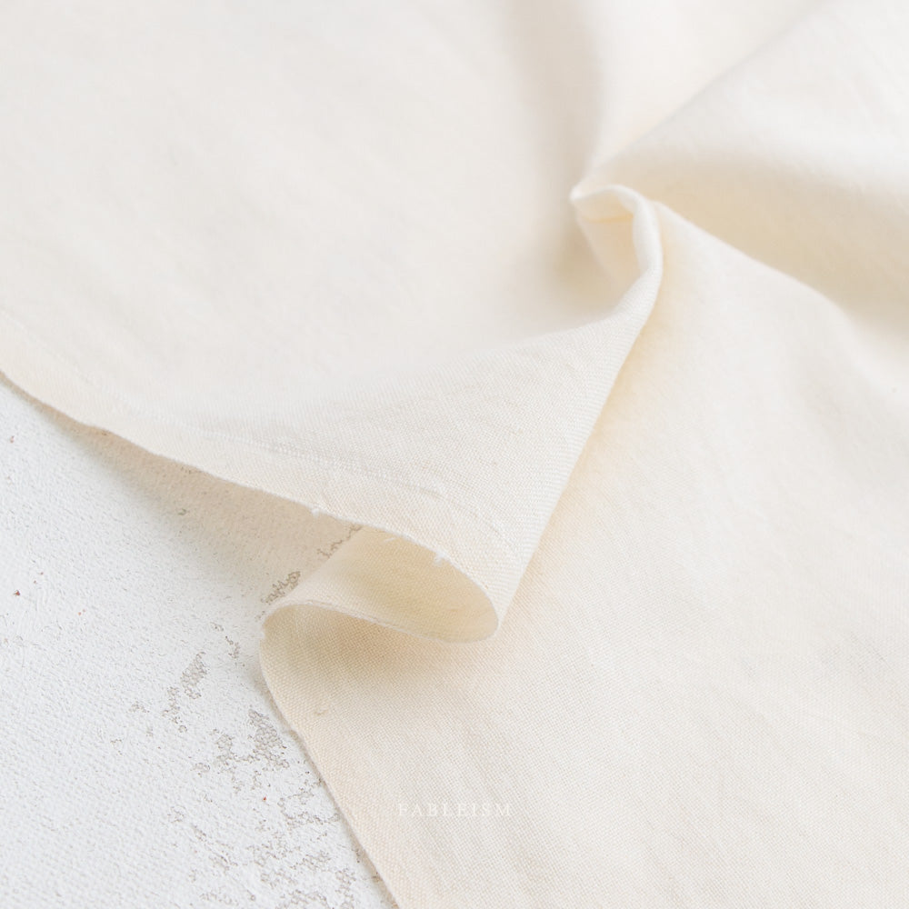 A close-up of Watercolor Wovens in Candlelight for Fableism: off-white, slightly textured 100% cotton with unfinished edges, draped on a speckled surface—ideal for patchwork or Watercolor Wovens projects.
