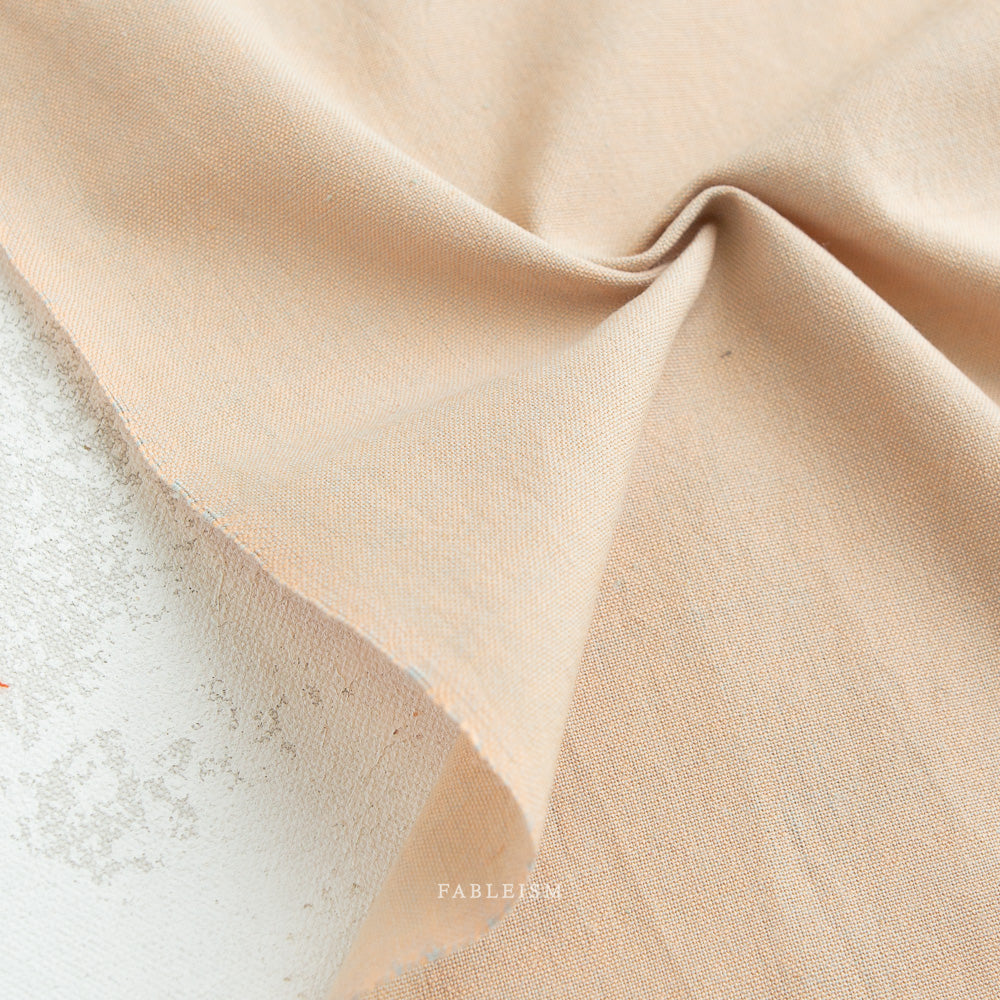 A close-up of Watercolor Wovens in Champagne for Fableism, 100% cotton with a visible weave, lightly wrinkled and folded on a textured white background—ideal for patchwork or quilting projects. The word FABLEISM appears faintly on the fabric.