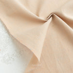A close-up of Watercolor Wovens in Champagne for Fableism, 100% cotton with a visible weave, lightly wrinkled and folded on a textured white background—ideal for patchwork or quilting projects. The word FABLEISM appears faintly on the fabric.