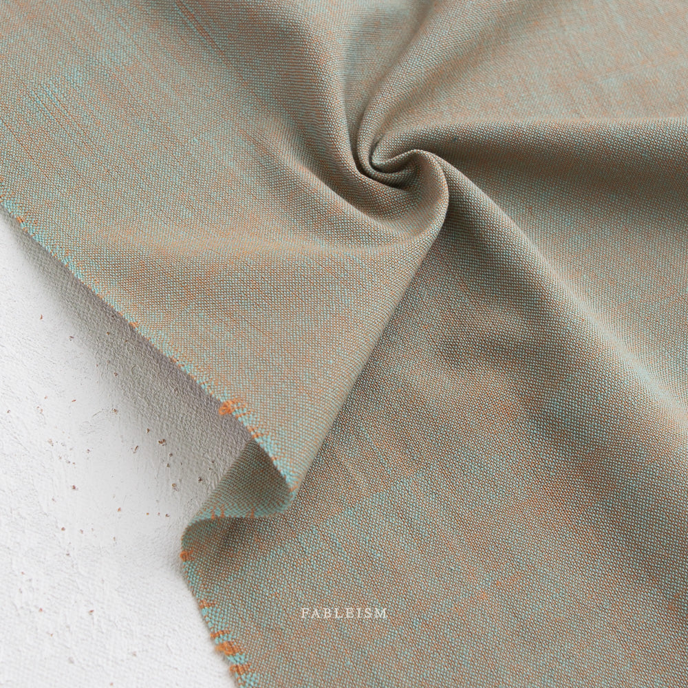 A close-up of Watercolor Wovens in Patina for Fableism, a textured light brown and blue 100% cotton fabric shown swirled on a white surface, ideal for patchwork. "FABELISM" is printed beneath the fabric.