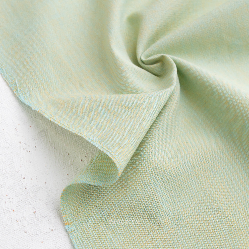 A close-up of Watercolor Wovens in Meadow for Fableism shows soft, lightweight, light green 100% cotton fabric with a subtle texture, draped and twisted on a white surface—ideal for patchwork or Watercolor Wovens projects.