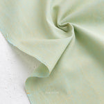 A close-up of Watercolor Wovens in Meadow for Fableism shows soft, lightweight, light green 100% cotton fabric with a subtle texture, draped and twisted on a white surface—ideal for patchwork or Watercolor Wovens projects.